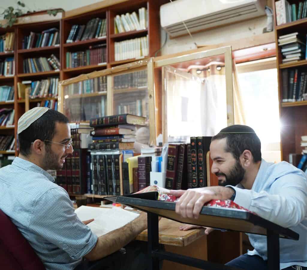 שני גברים חובשי כיפה יושבים אחד מול השני עם ספר תורה ומחייכים